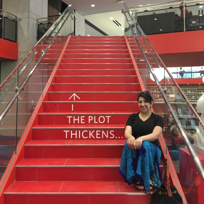 Emily is sitting on a red staircase at Boston Public Library. The text "The plot thickens..." is written on the stairs.