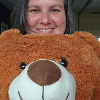 My smiling face framed by brown hair is stacked on top of a reddish-brown stuffed teddy bear's smiling face. We are story time bros.
