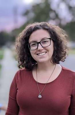 Photo of a white woman with curly hair and black glasses smiling at the camera