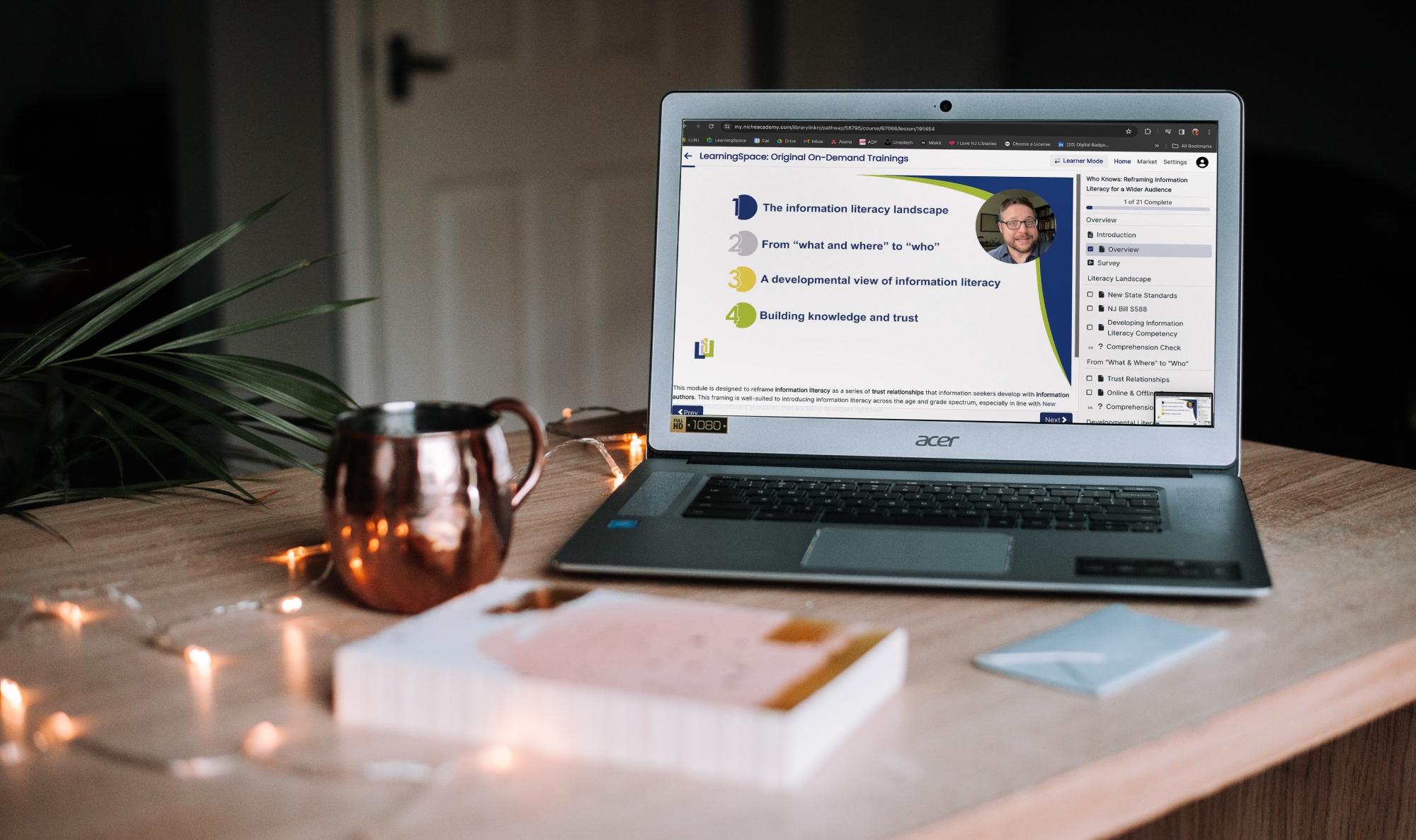 A laptop sits open with the presentation playing on it. The scene is a cozy home with a coffee mug and book presented on the table, beside the laptop.