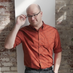 Jon stands against a brick wall, wearing black slacks and a brown dress shirt. He is smiling while reaching up to adjust his glasses.