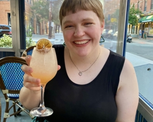 Melody is sitting a table, wearing black dress and green triangle earrings, holding a cocktail and smiling.