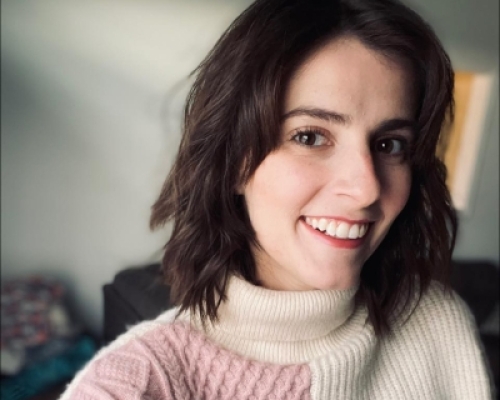 Woman with brown hair wearing a pink and white sweater in front of a grey background.