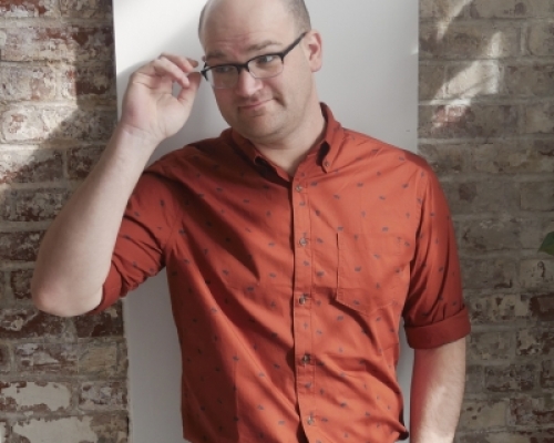 Jon stands against a brick wall, wearing black slacks and a brown dress shirt. He is smiling while reaching up to adjust his glasses.