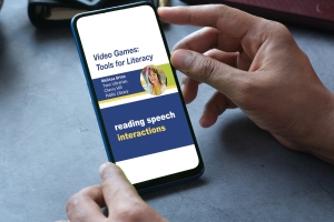 A person holds a smartphone as a clip is displayed from the webinar Video Games: Tools for Literacy with presenter Melissa Brinn.