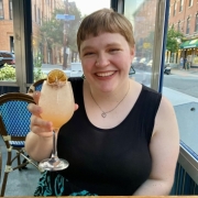 Melody is sitting a table, wearing black dress and green triangle earrings, holding a cocktail and smiling.